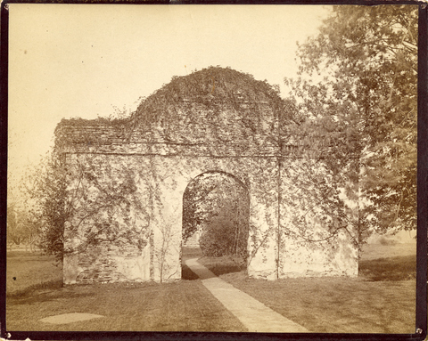 Sepia-toned black-and-white photograph of a stone arch covered in ivy with a footpath running through the opening.