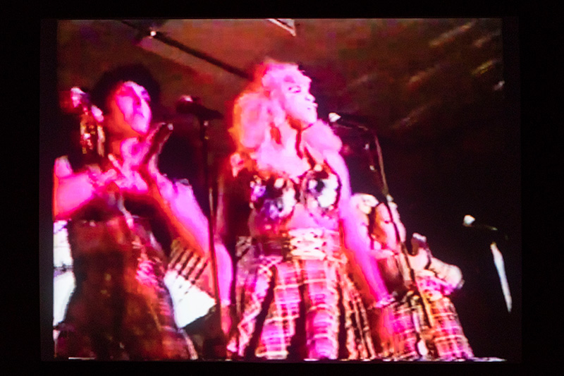 Performers front of stage, bathed in pink light, wearing plaid skirts and metal bustiers