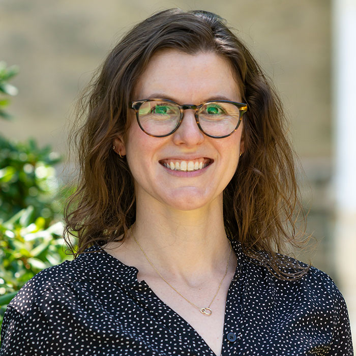 Headshot of Jess Libow, smiling in tortoiseshell pattern glasses against a soft focus background of the Haverford College campus