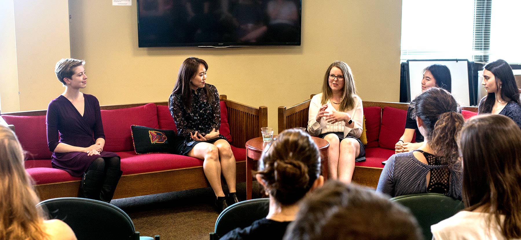 Three women sit on couches adressing a group of students