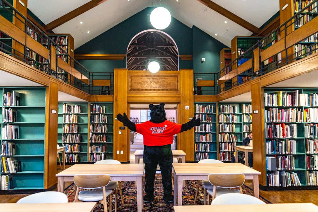 Black Squirrel mascot standing among a wing in a library. There are a multitude of shelves of books behind it.