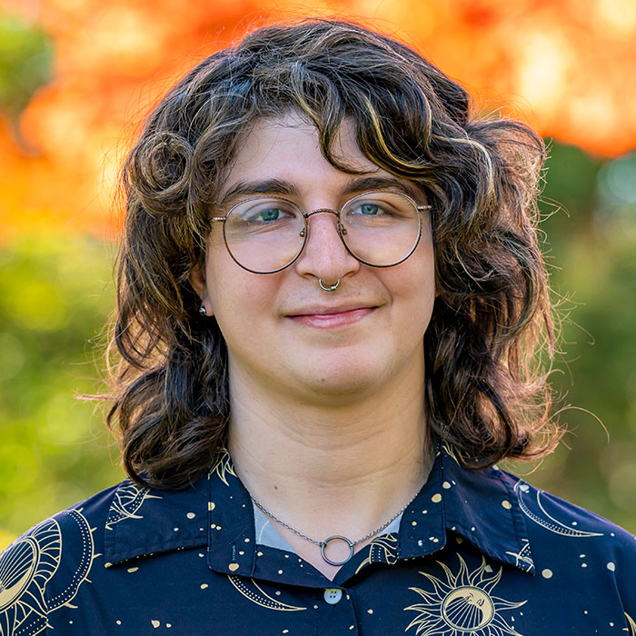 Headshot of June Scafiro, wearing glasses, earrings, and a white sweater with the collar of a white button down with navy blue astronomy print visible