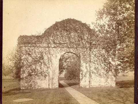 Sepia-toned black-and-white photograph of a stone arch covered in ivy with a footpath running through the opening.