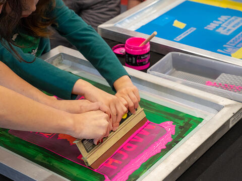 Two sets of hands drag a squeegee and pink ink across a screen printing screen. 