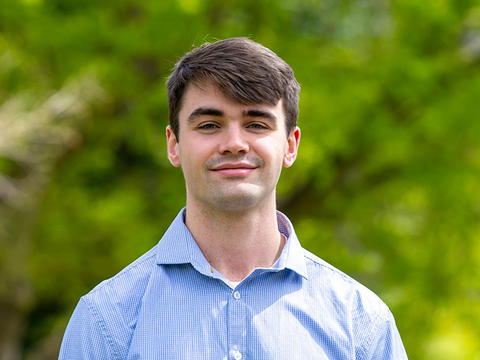 Gabe Jones-Thomson smiling for the camera in a light blue button down shirt against a soft focus backdrop of Haverford College foliage