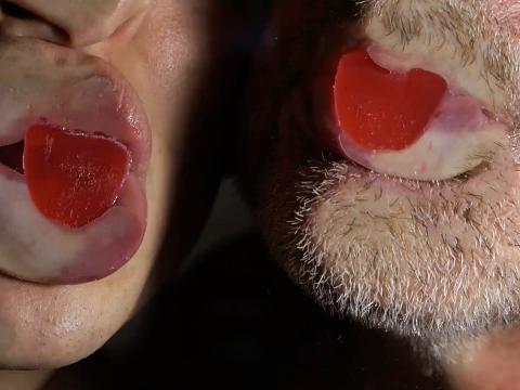 The lower half of two faces, one bearded, smashed against glass with red candy in the mouths