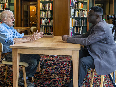 Two men sit at a table in a library setting while filming a documentary. 