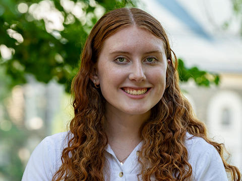 Abigail Pruszinski smiles in a portrait taken on Haverford's campus. Behind her are out-of-focus trees and a campus building. 