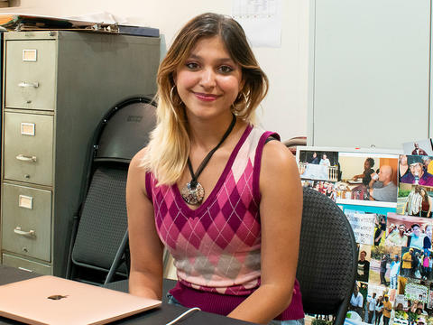 Anjali Agarwal sits at a desk at her internship near filing cabinets and a collage of photographs. She wears a pink argyle sweater and a large necklace. 