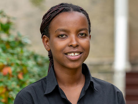 Ghislaine Niyodusenga looks at the camera in a portrait. She wears a black button up shirt and is pictured in front of a building on Haverford's campus. 