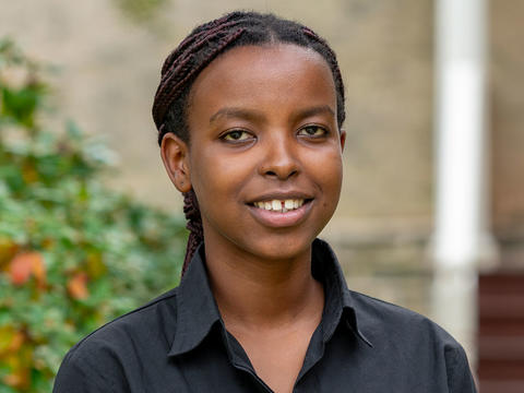 Ghislaine Niyodusenga looks at the camera in a portrait. She wears a black button up shirt and is pictured in front of a building on Haverford's campus. 