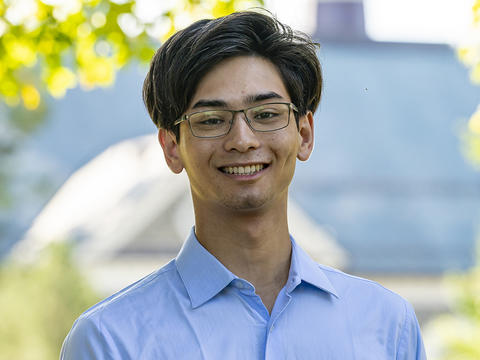 Brendan Holleck is pictured in a portrait on Haverford's campus. He wears glasses and a blue button-up shirt. Behind him, the background is out of focus. 