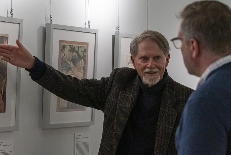 Arnold Satterwait, wearing a black turtleneck and dark jacket, gestured to a framed Japanese print in a gallery.