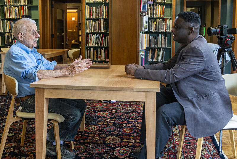 Two men sit at a table in a library setting while filming a documentary. 