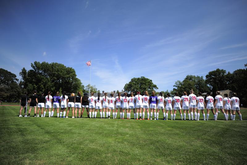 The women's soccer team stands in a line in their uniforms with the American flag waving in the background