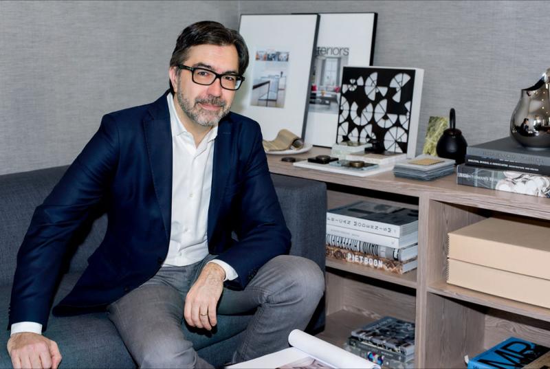 Jean Gabriel Neukomm '90, wearing glasses and a blue blazer, sits on a low gray couch next to a bookshelf that's full of art books and prints