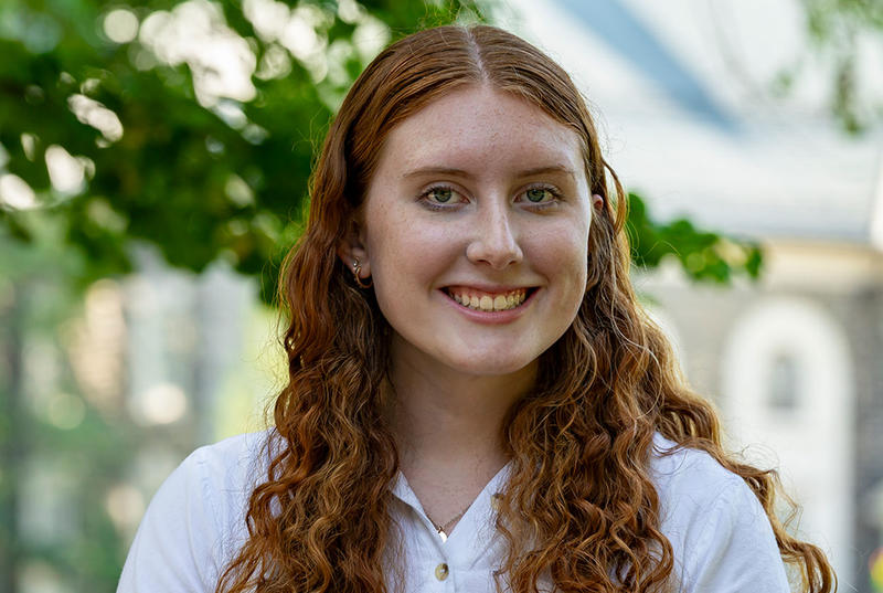 Abigail Pruszinski smiles in a portrait taken on Haverford's campus. Behind her are out-of-focus trees and a campus building. 