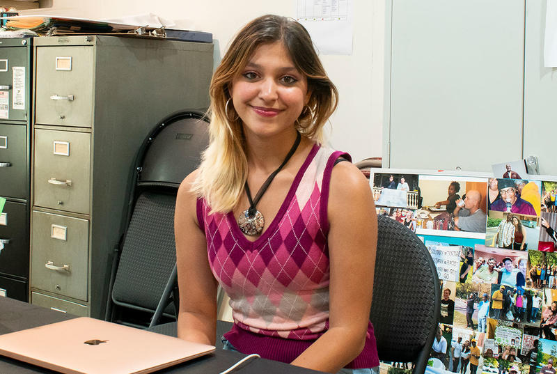 Anjali Agarwal sits at a desk at her internship near filing cabinets and a collage of photographs. She wears a pink argyle sweater and a large necklace. 