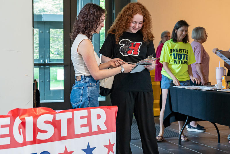 Two students look at a clipboard as they register to vote in the GIAC. One is wearing a black Haverford t-shirt with the black squirrel on it. 