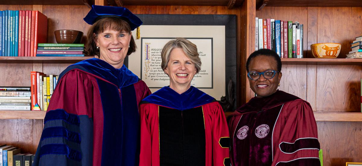 Three women in academic robes stand together in front of bookcases