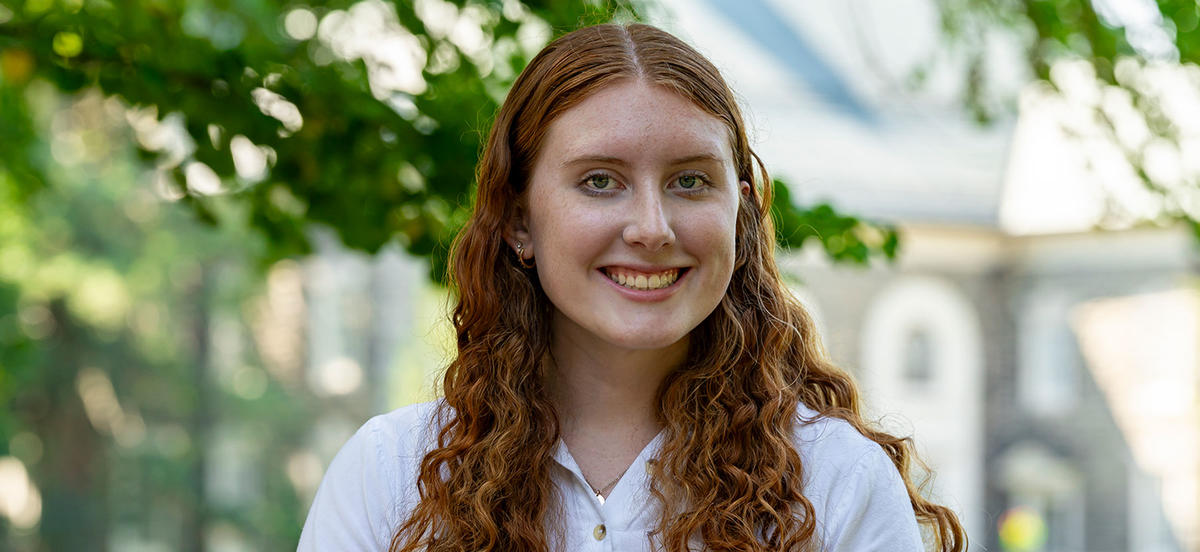 Abigail Pruszinski smiles in a portrait taken on Haverford's campus. Behind her are out-of-focus trees and a campus building. 