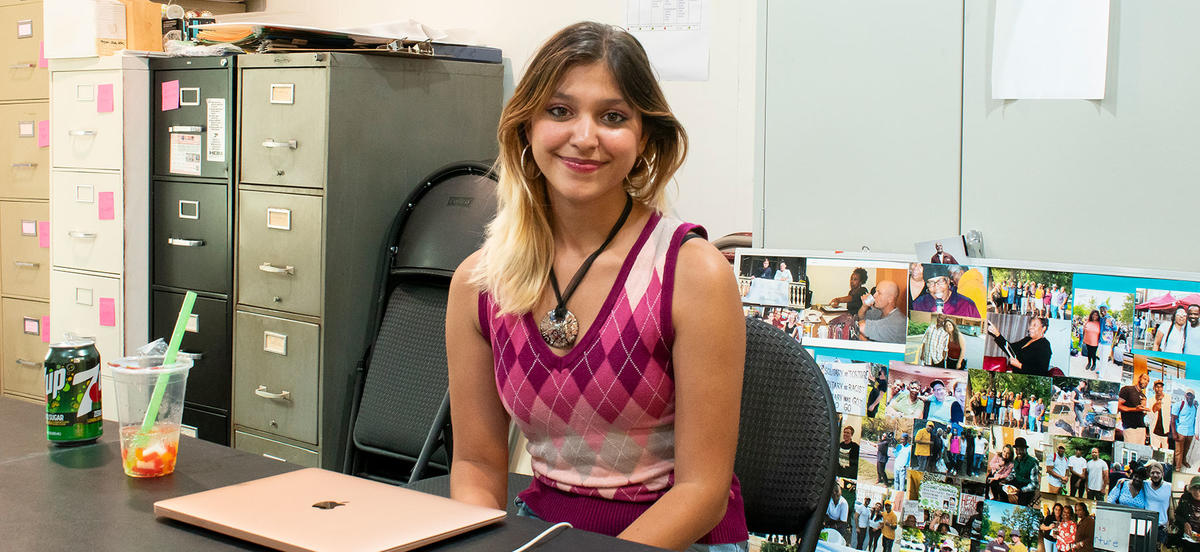 Anjali Agarwal sits at a desk at her internship near filing cabinets and a collage of photographs. She wears a pink argyle sweater and a large necklace. 